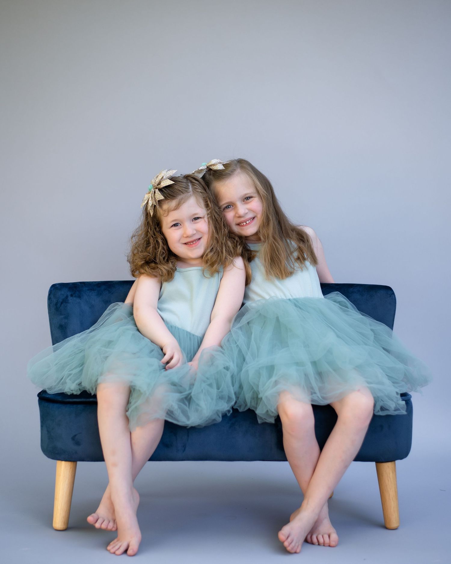 Two girls in matching turquoise tulle dresses sit on navy blue bench during studio portrait session.