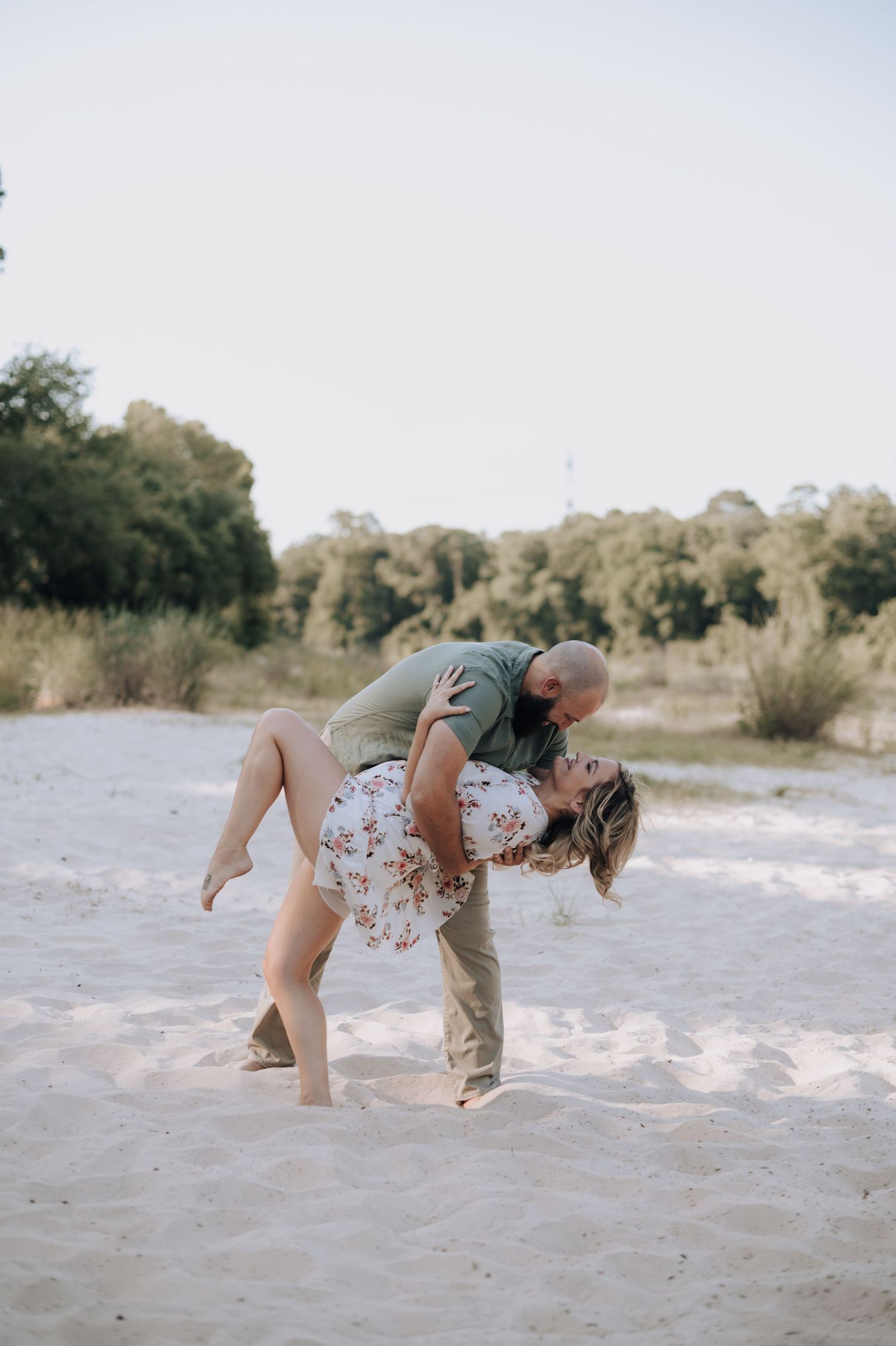 A romantic dip pose captured on a sandy beach with trees in the background.
