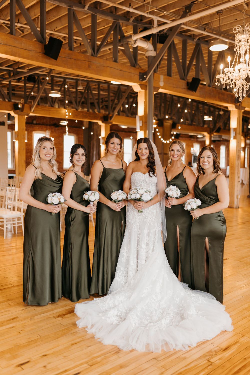 Bride in lace gown poses with five bridesmaids in olive green satin dresses holding white bouquets.
