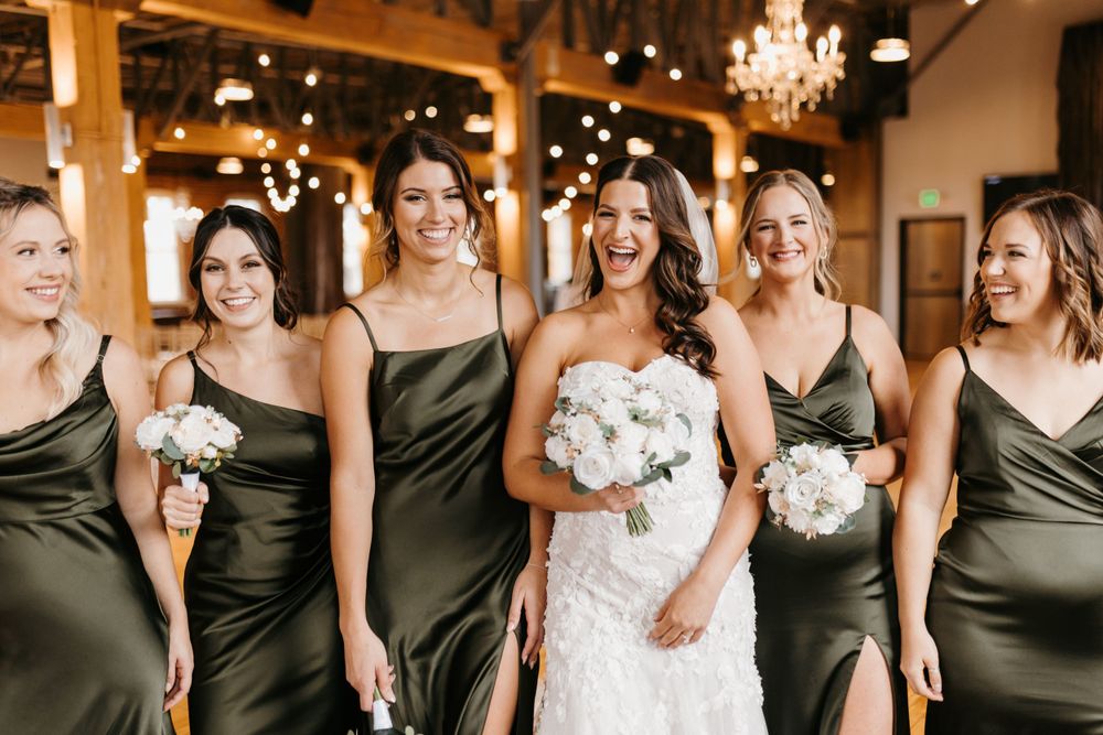 Bride in lace gown laughs with bridesmaids in olive green satin dresses holding white bouquets.