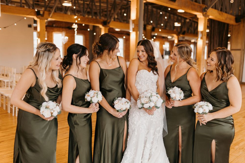 Bridesmaids in olive green satin gowns laugh with bride in lace dress holding white floral bouquets.