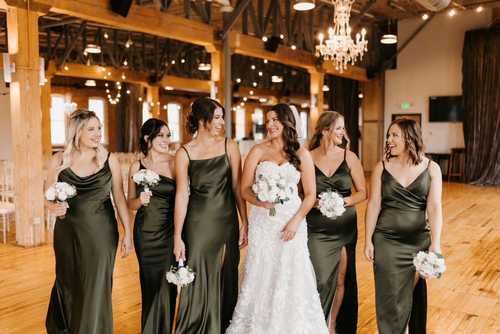 Bride in strapless lace gown laughs with bridesmaids in olive satin dresses in rustic barn venue.