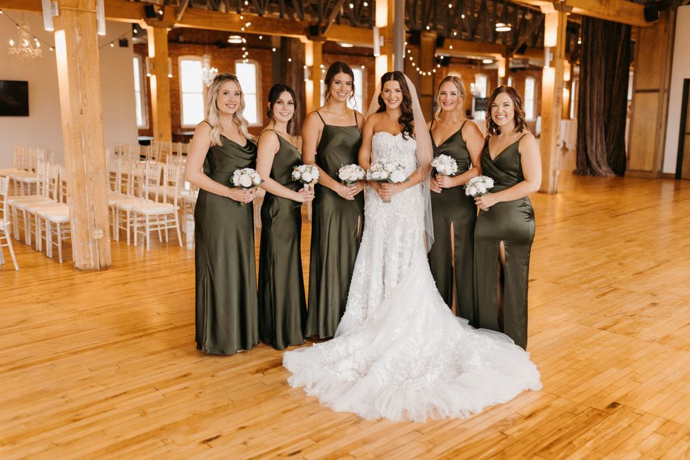 Bride in lace wedding gown stands with bridesmaids in olive satin dresses in rustic barn venue.