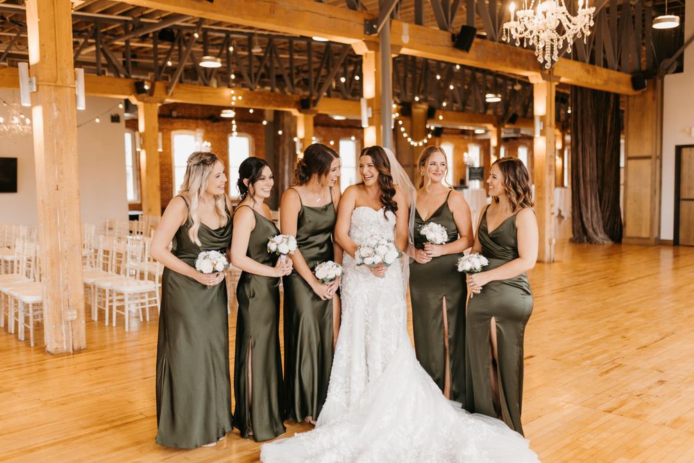 Bride in lace gown walks with bridesmaids in olive green satin dresses in elegant barn venue.