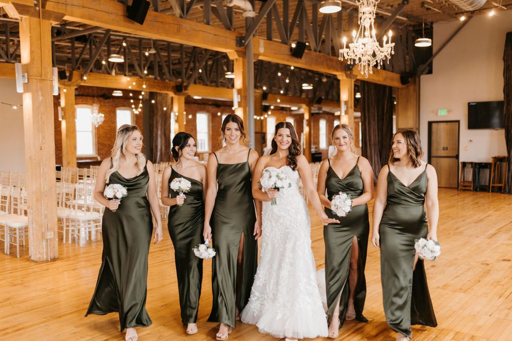 Bride and bridesmaids in olive green satin gowns walk together in chandelier-lit rustic barn venue.
