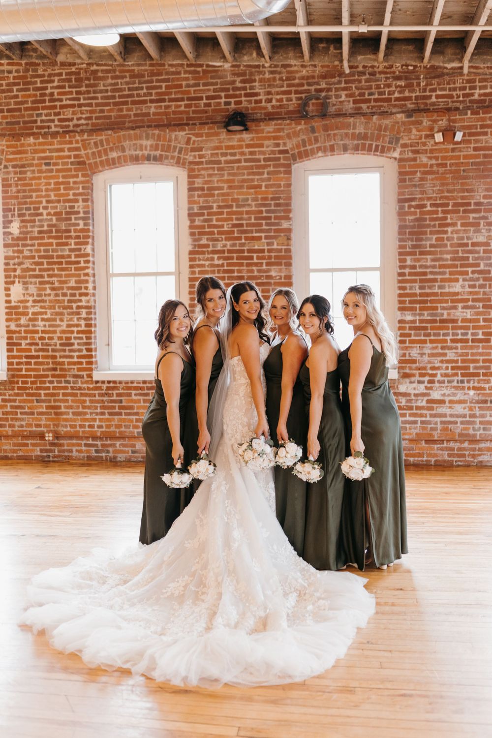 Bride in lace gown with bridesmaids in olive green dresses holding bouquets in rustic brick venue.