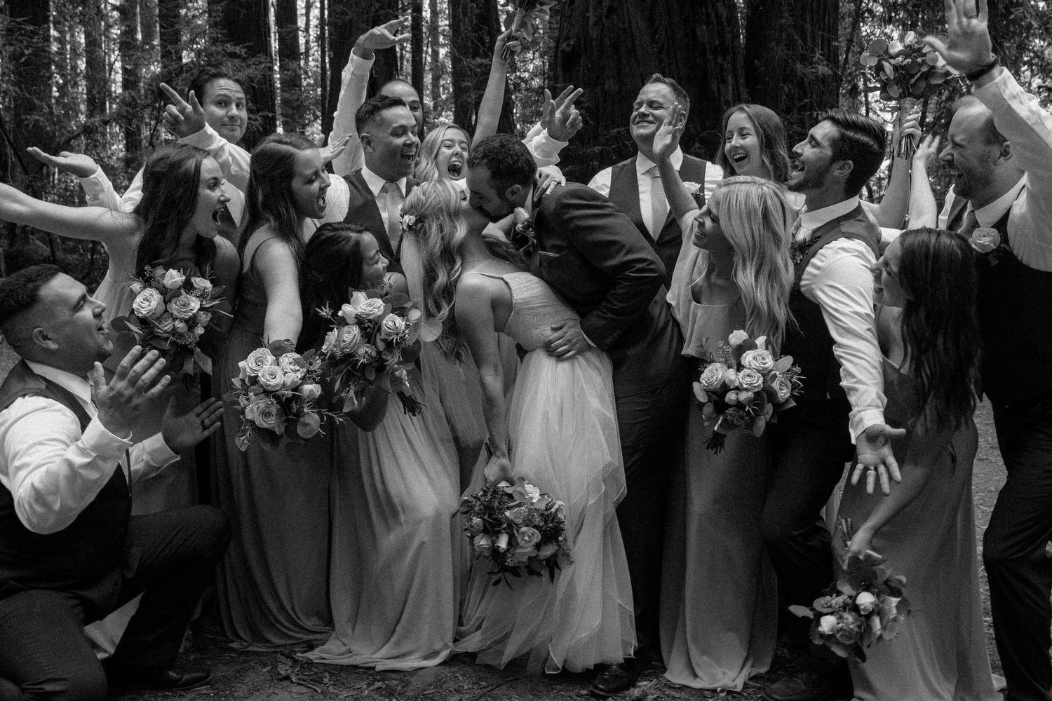 Black and white wedding party photo with bridesmaids and groomsmen celebrating in a forest setting.