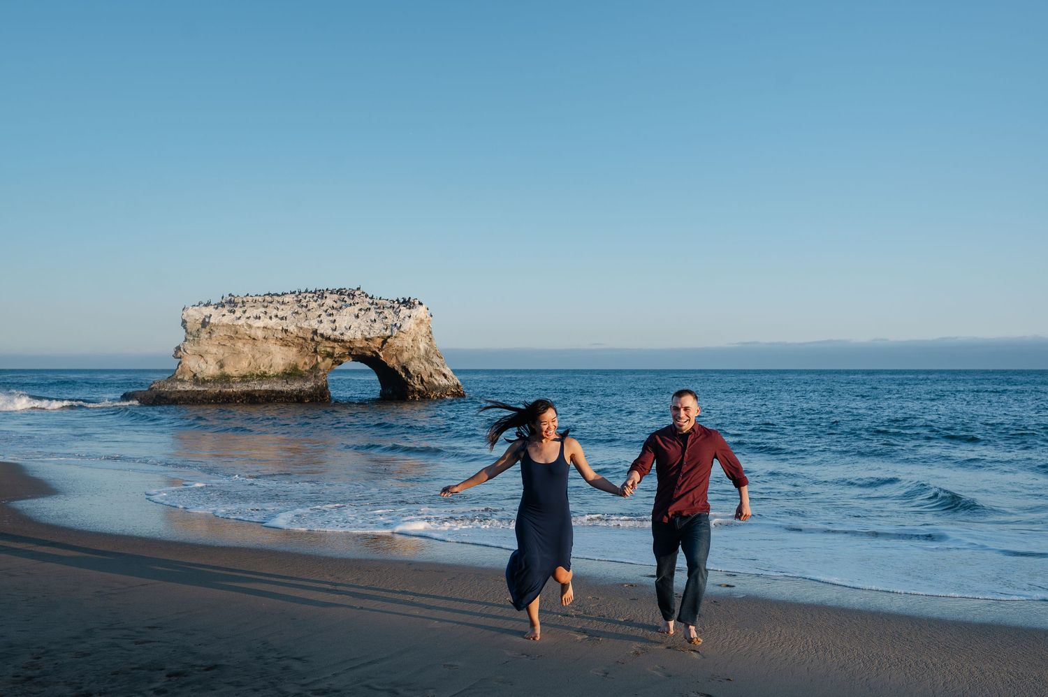 Two people run playfully along the shoreline with a natural rock arch visible in the ocean behind them.