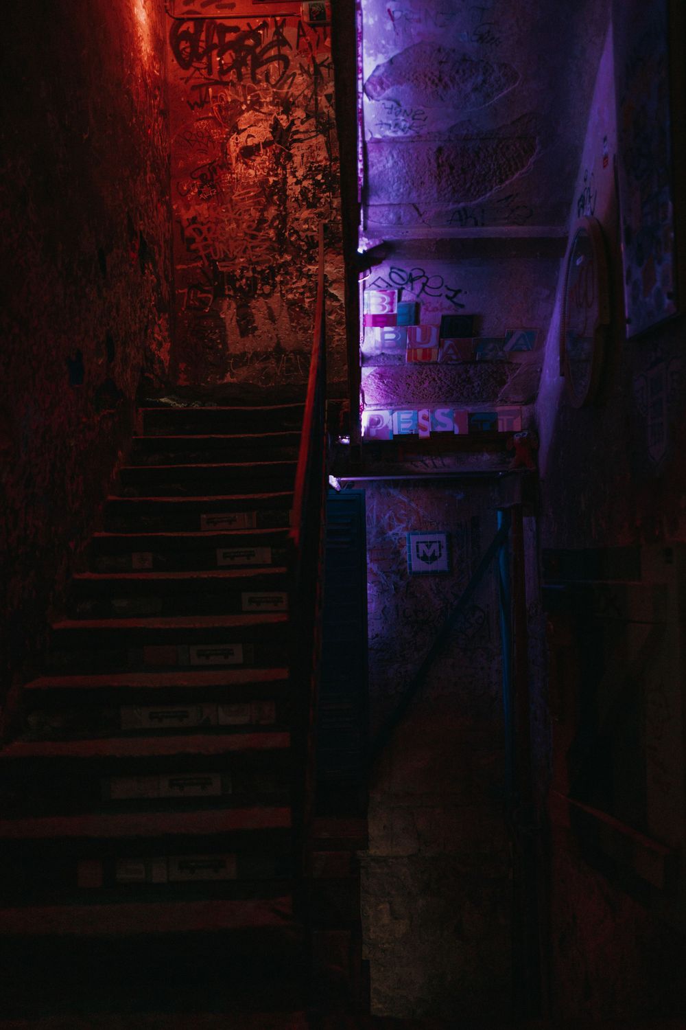Dark stairwell illuminated by eerie red and purple lighting with graffiti covered walls.