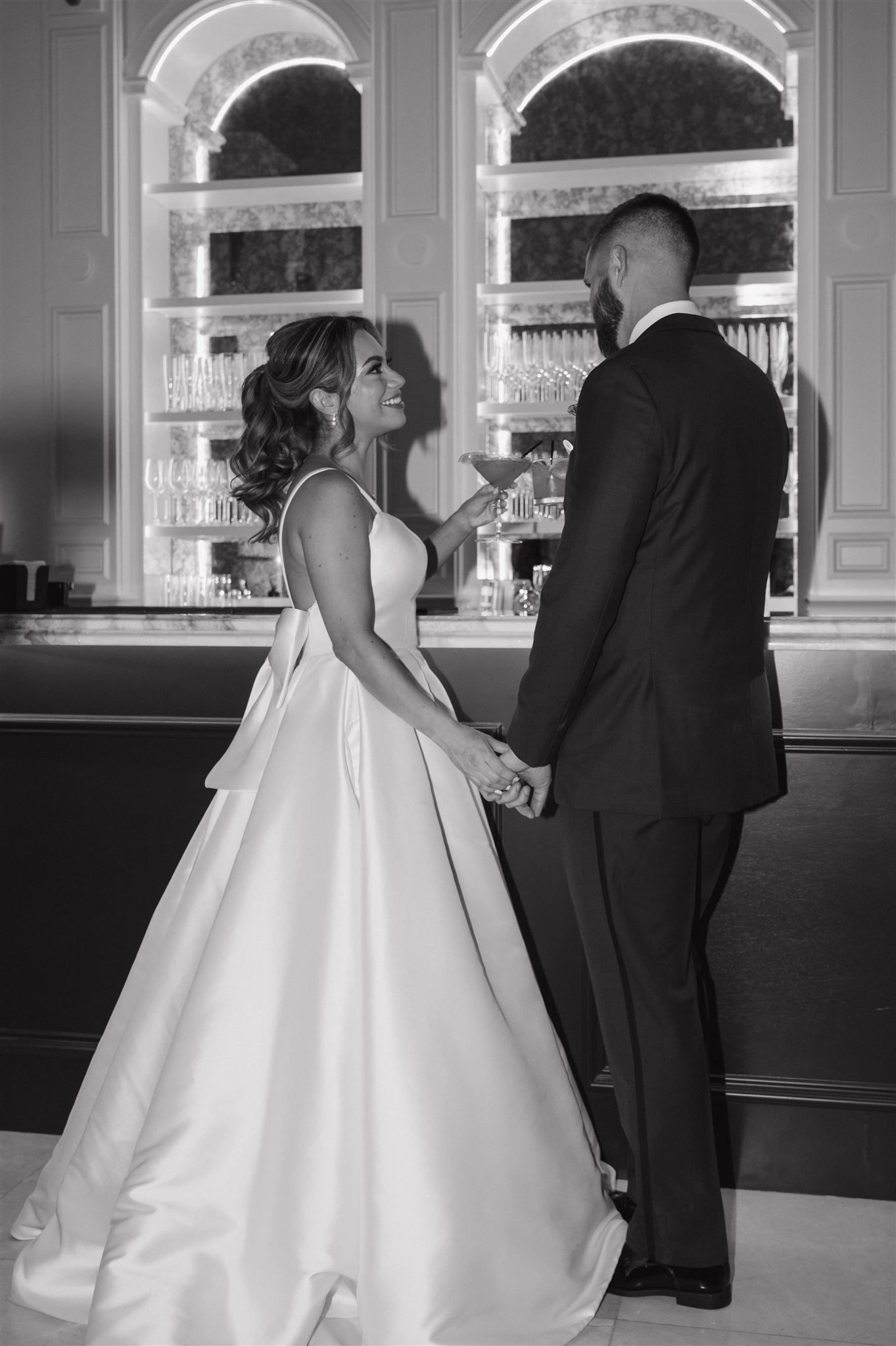 A couple at a formal bar in wedding attire, with ornate arched windows and cabinets in the elegant background.