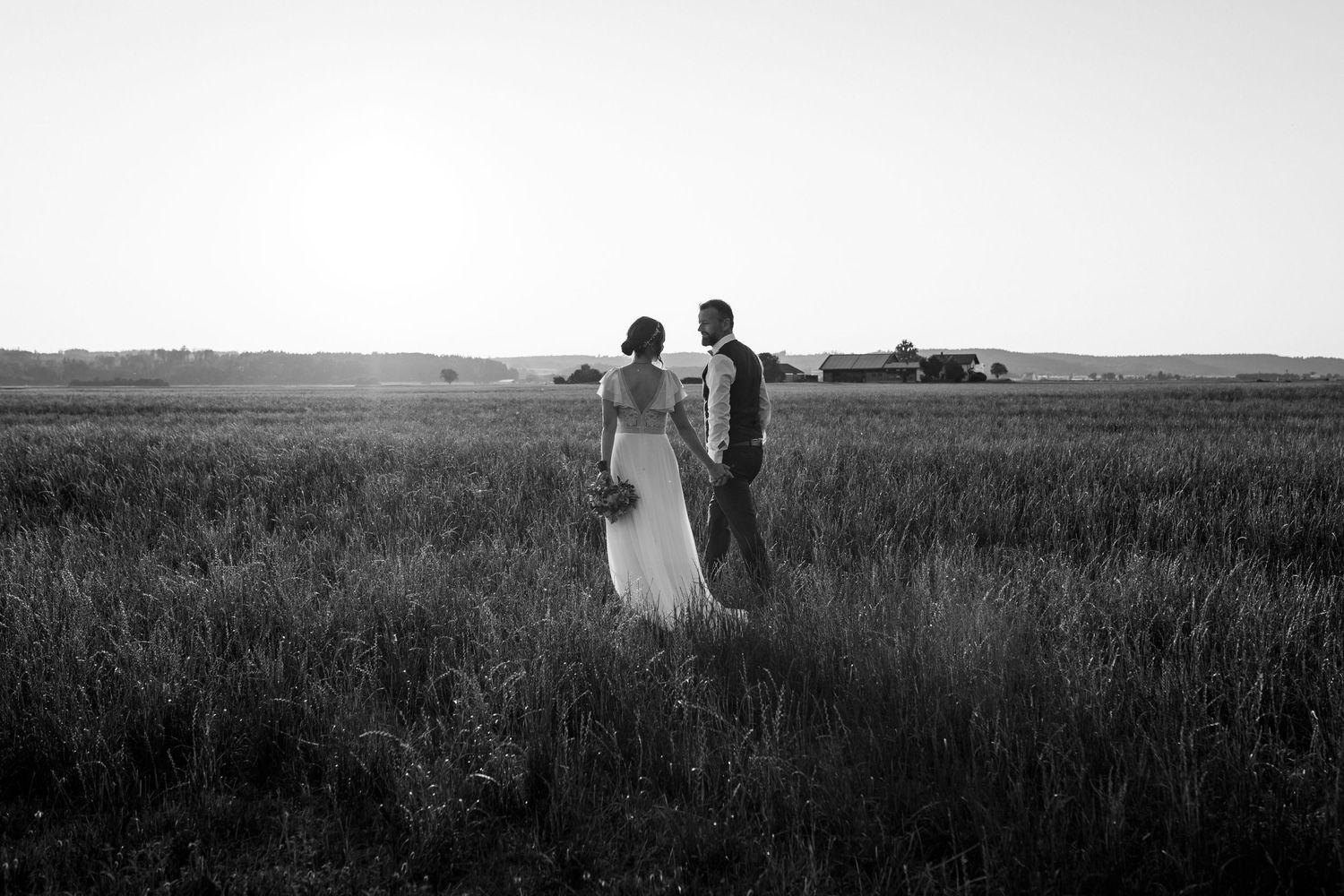 Ein Paar in Hochzeitskleidung hält sich an den Händen, während es bei Sonnenuntergang in Schwarz und Weiß durch ein rustikales Feld geht.