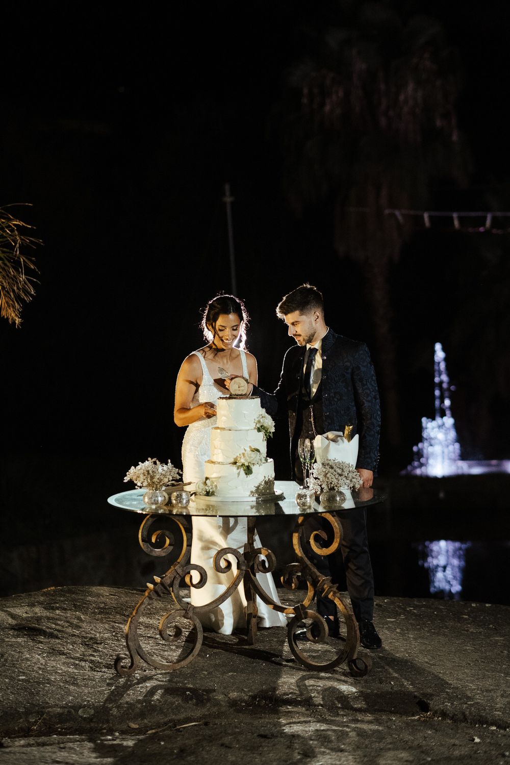 Casal corta o bolo de casamento junto à noite, com uma fonte iluminada ao fundo.