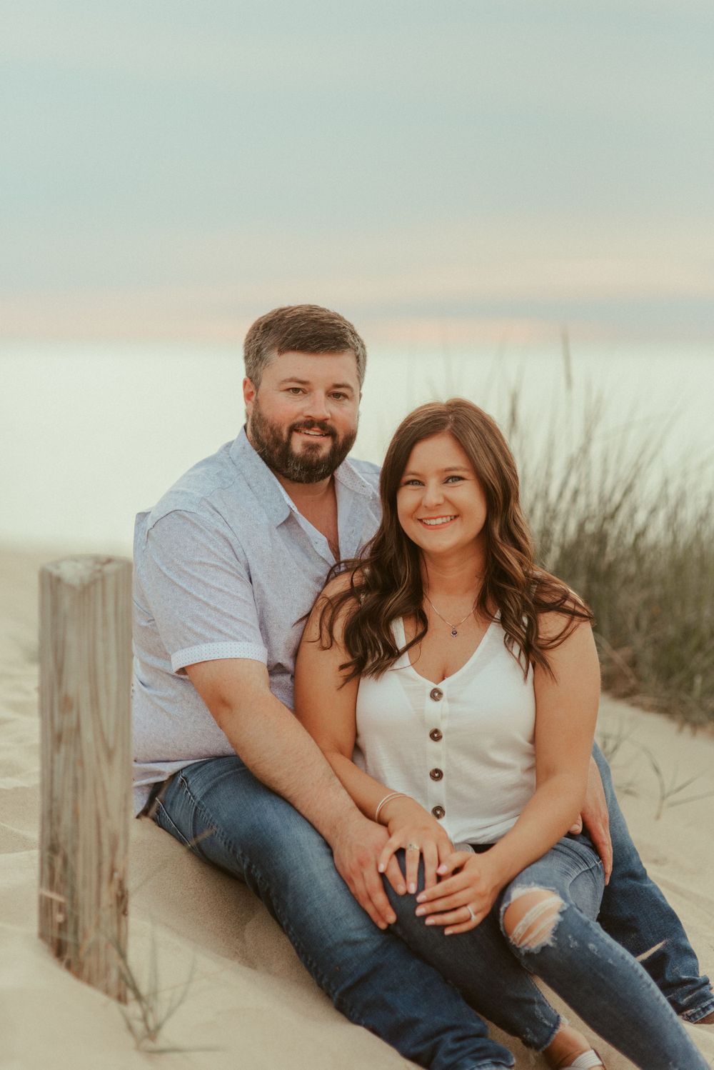 Couple sitting together on sandy beach dunes at sunset wearing casual denim and white outfits.