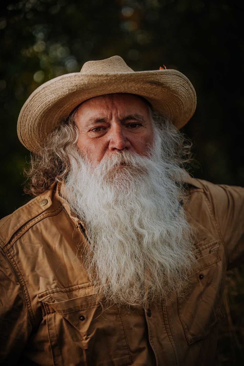 A rugged figure in tan workwear and straw hat stands against a dark natural backdrop.