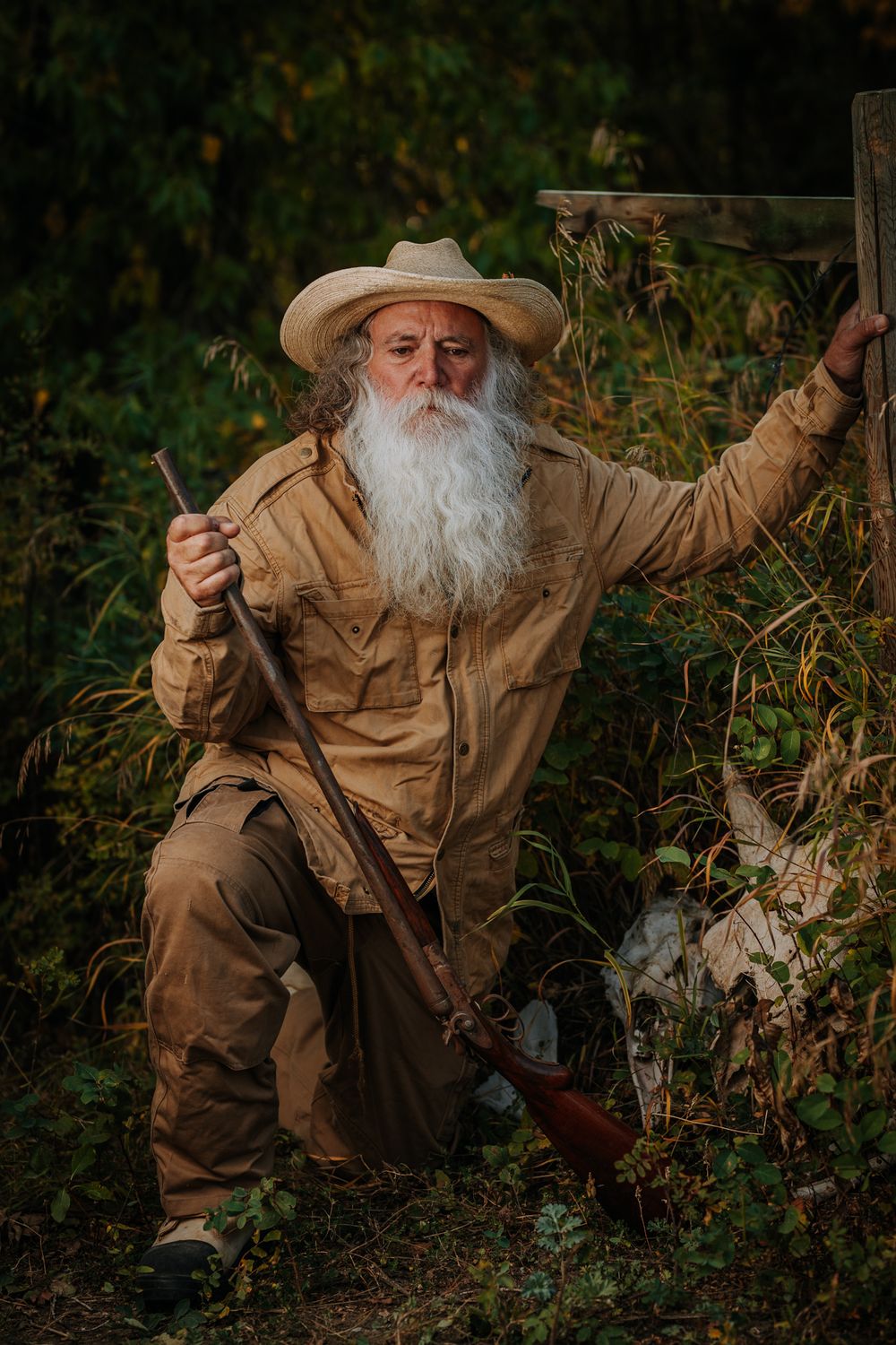 A rugged outdoorsman in canvas clothing crouches with vintage hunting gear in a natural setting.