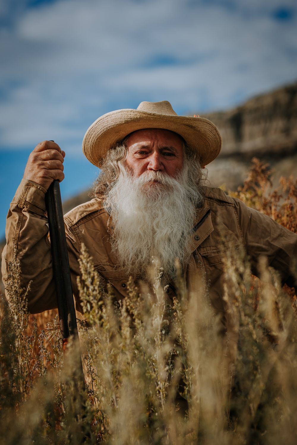 A figure in western attire stands among tall grass with rocky cliffs in the background.