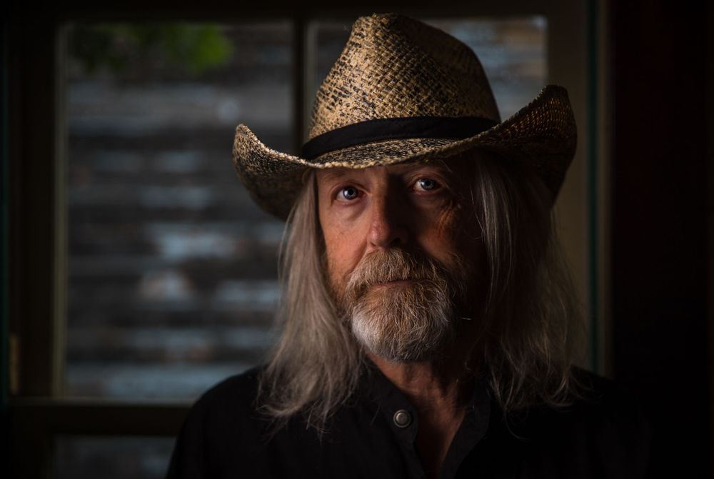Dramatic portrait featuring a weathered cowboy hat and long gray hair in moody lighting.