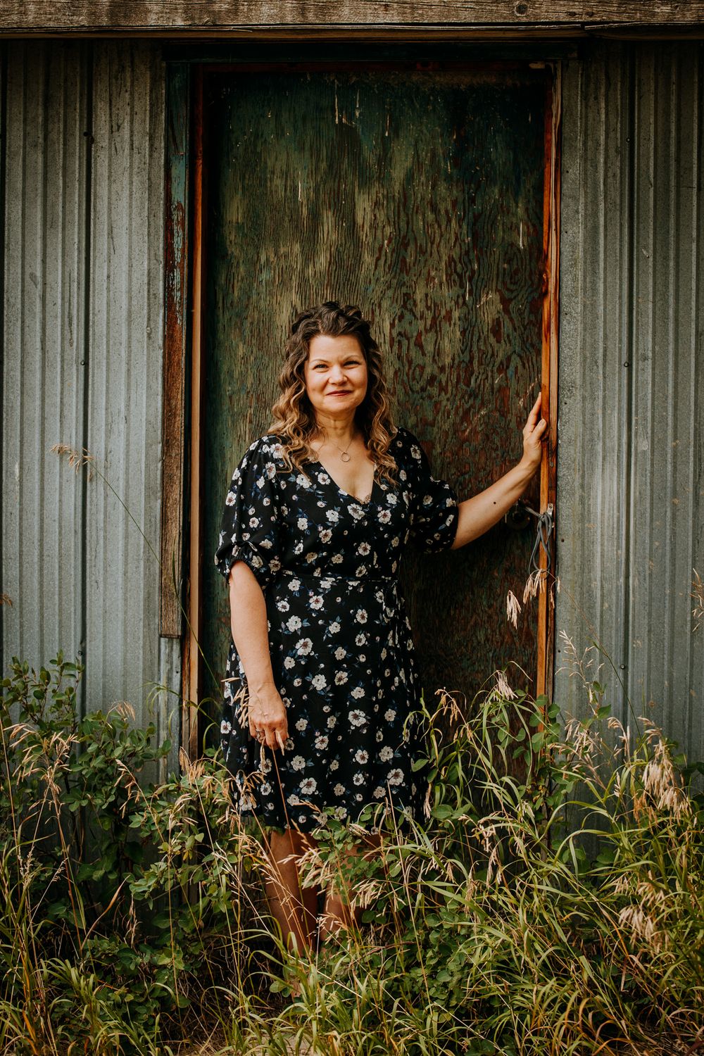 Portrait in front of weathered metal wall with tall grass and floral dress.