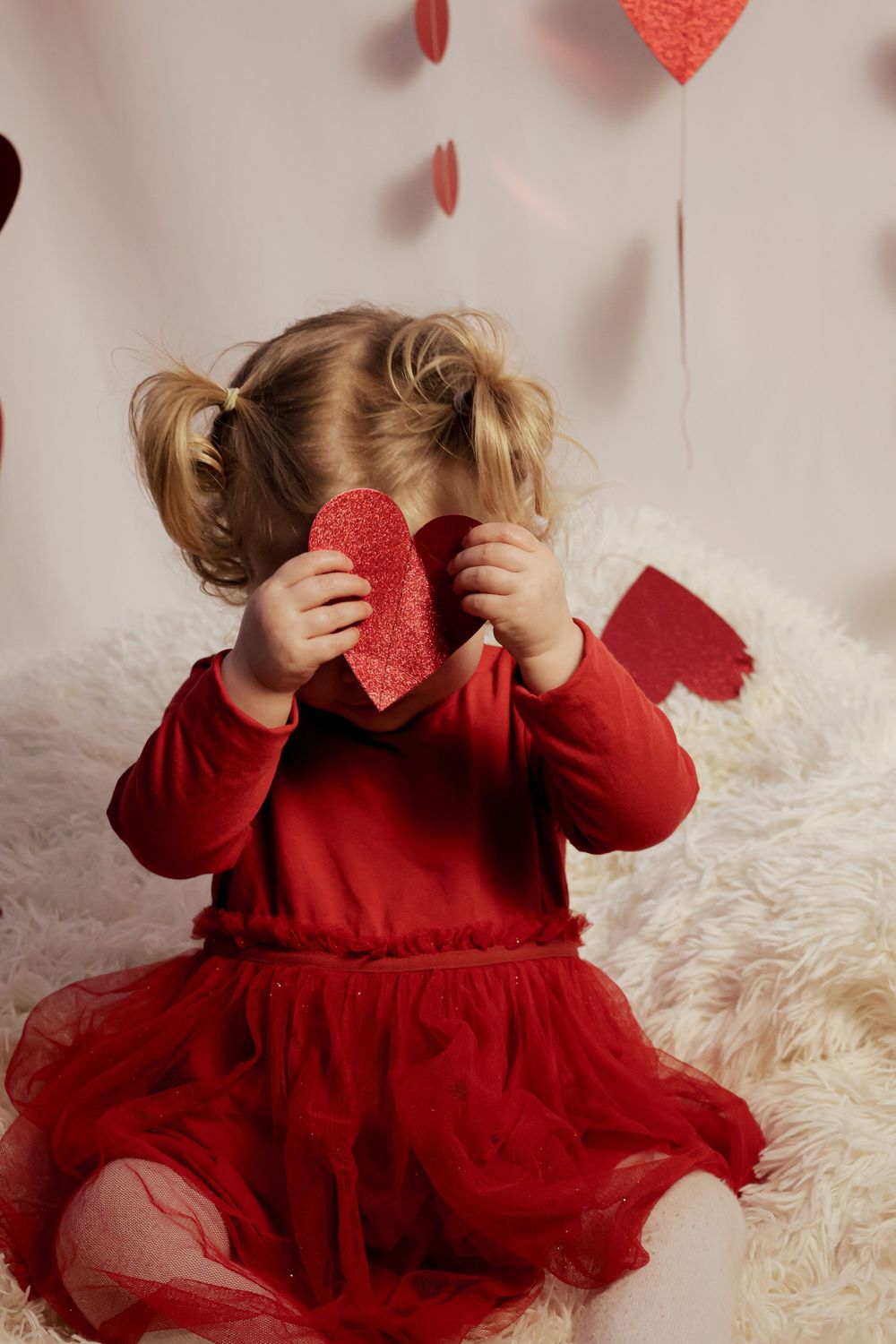 Toddler girl in red outfit holding a glittery red heart covering her face on Valentine's Day.