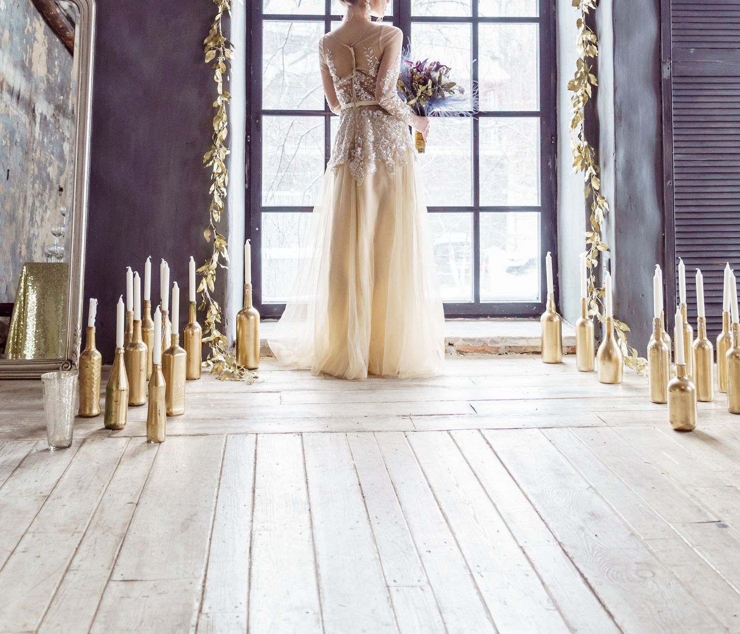 Elegant wedding dress displayed in a rustic room with gold candles and ornate window frame.