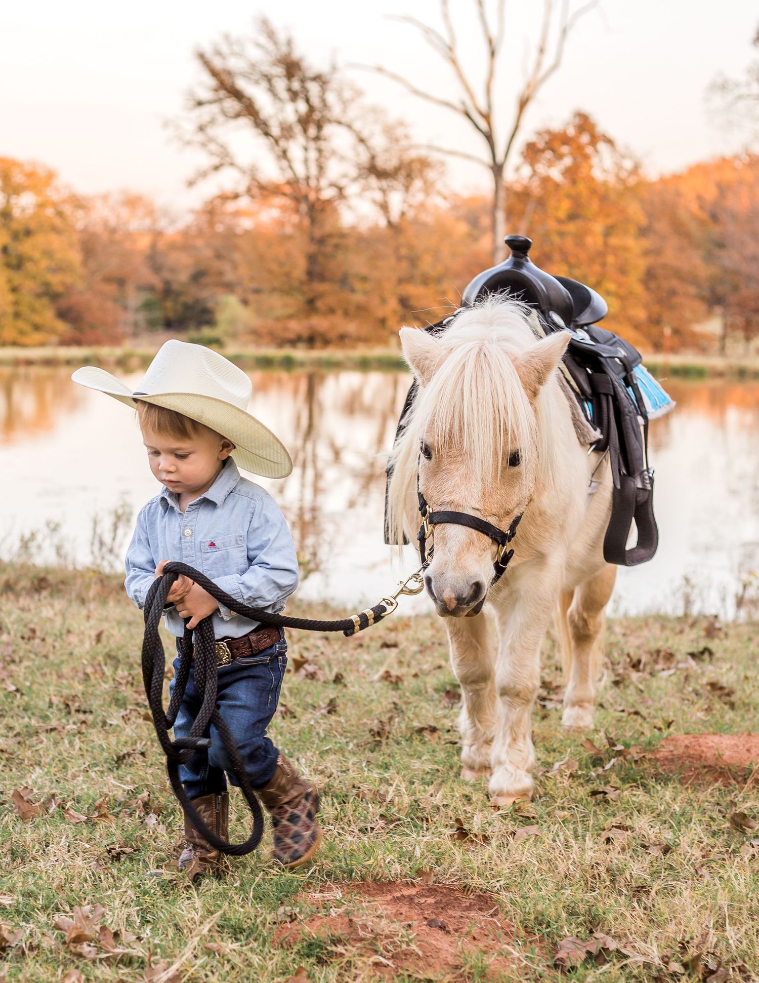 Young cowboy in hat leading a white pony by a lake during autumn.