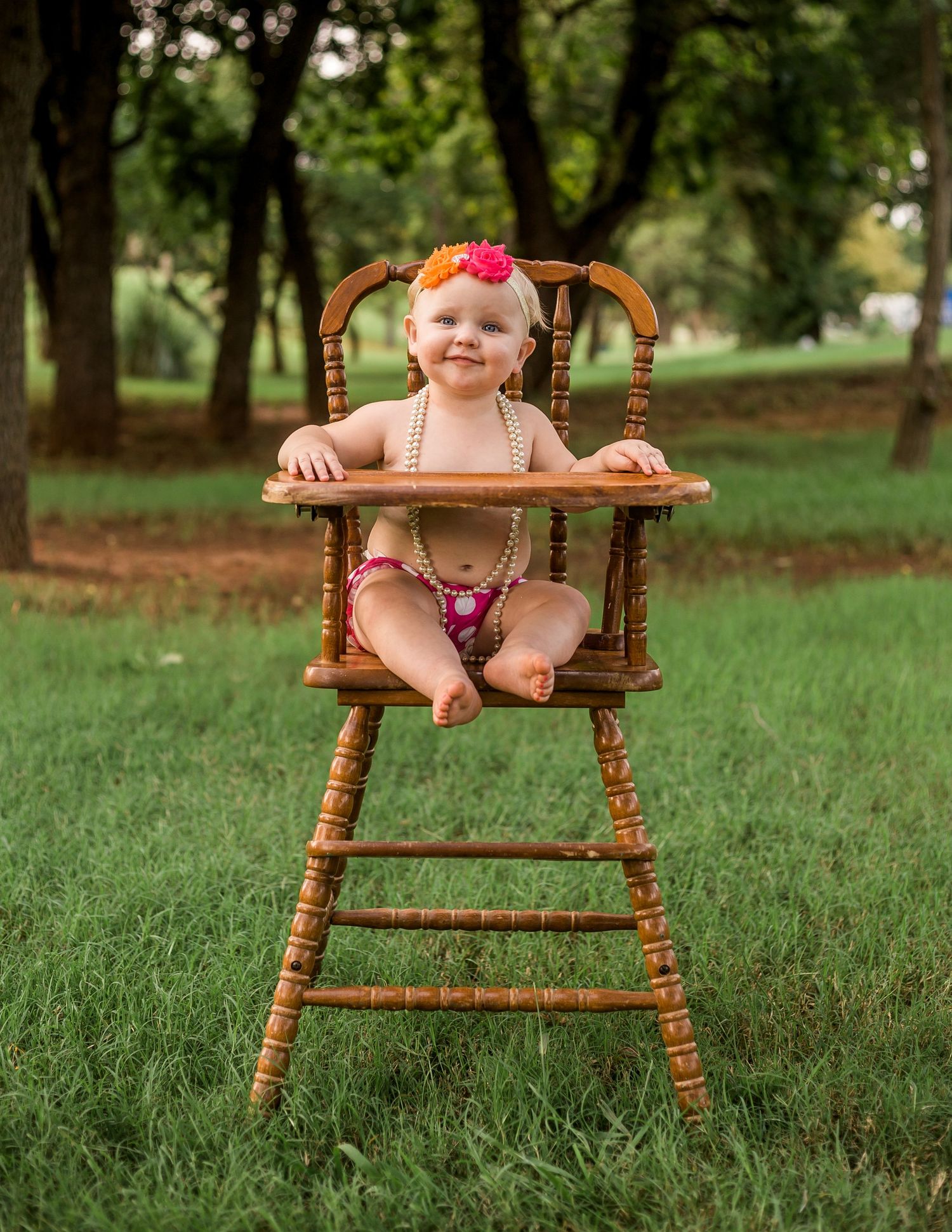 Baby wearing pink bow sits happily in vintage wooden high chair on green grass.