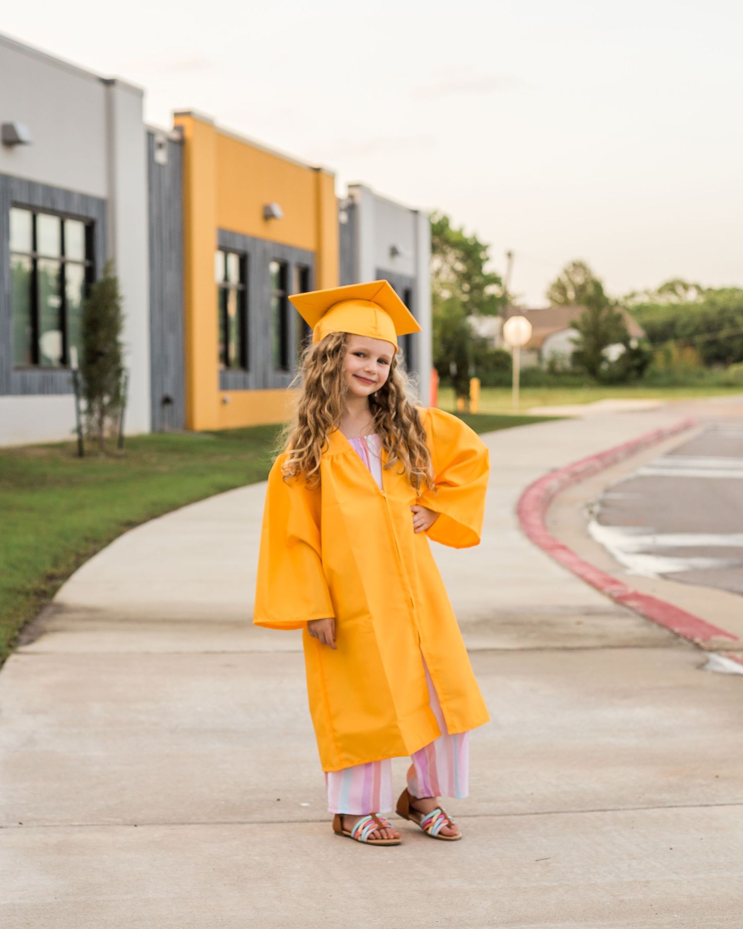 A bright orange graduation gown and cap worn outdoors near modern buildings.