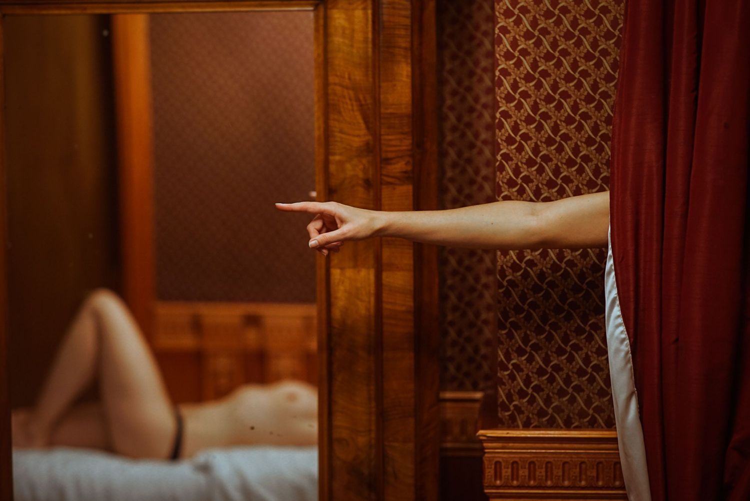 A woman is pointing at a mirror, drawing attention to the reflection of a naked woman lying on a bed.