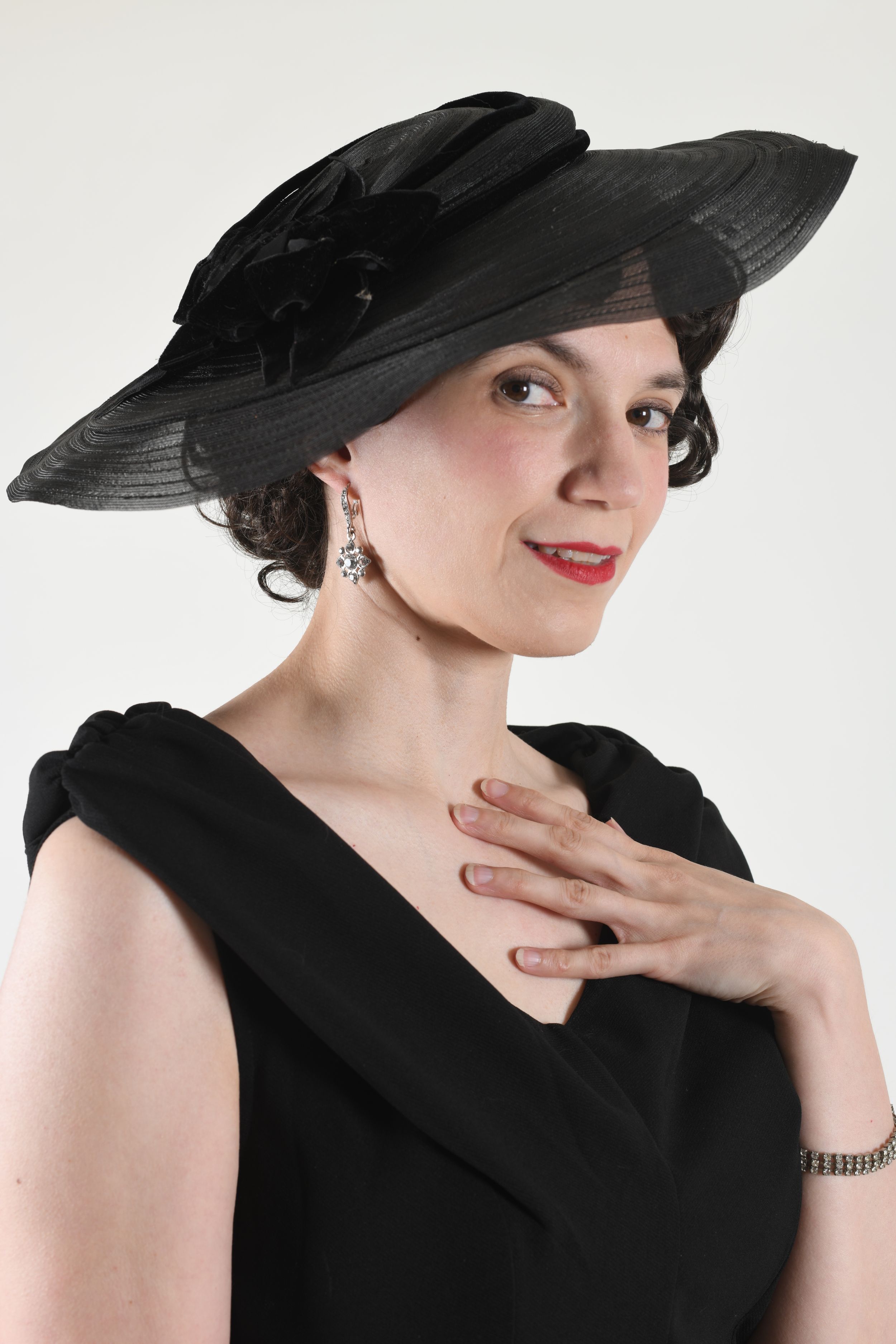 Elegant woman in vintage black dress and wide-brimmed hat posed against white background.