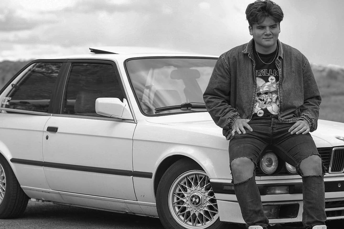 Classic BMW E30 car in black and white with rims and a person sitting on the hood.