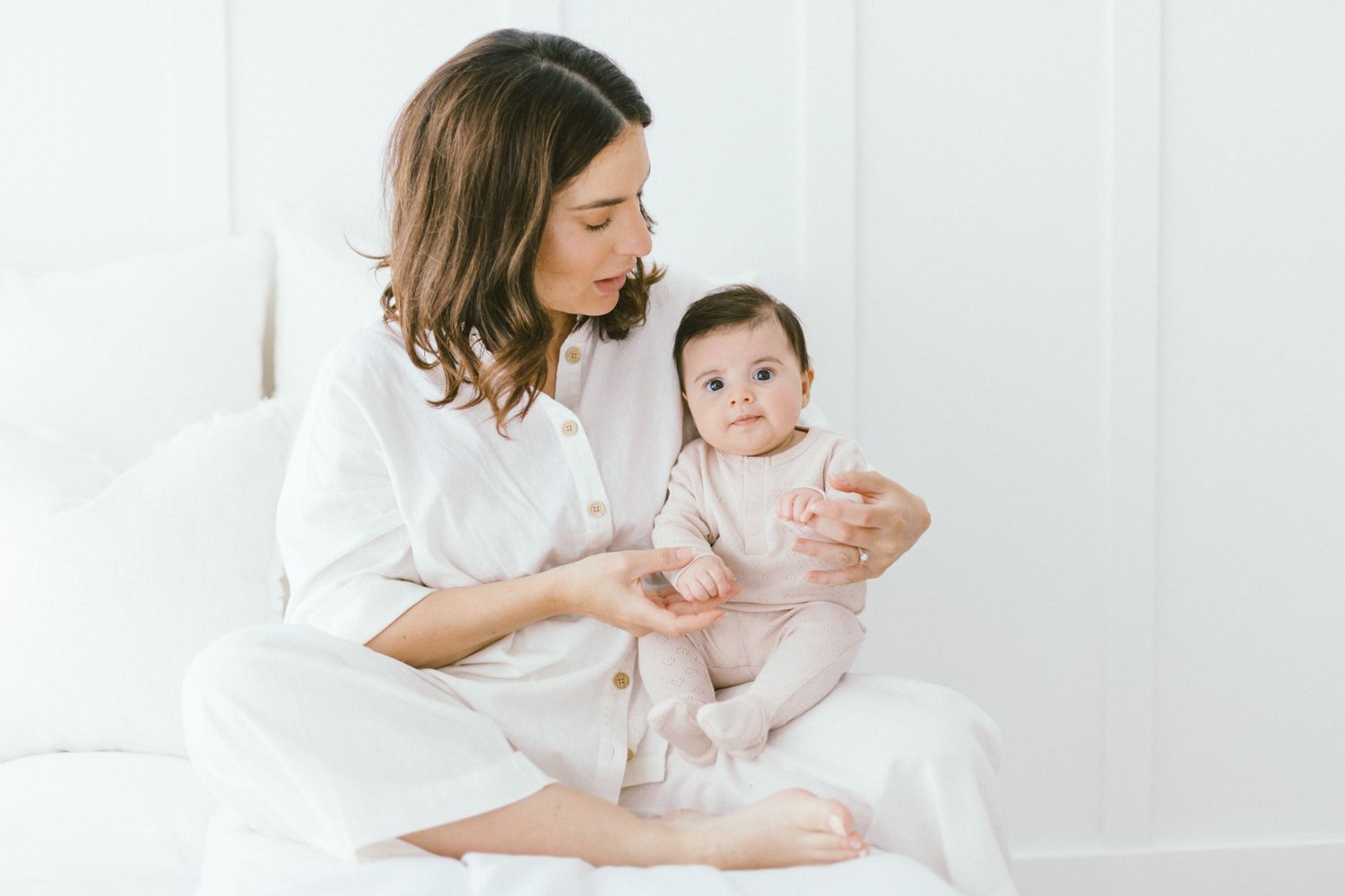 Intimate portrait of parent and baby in white clothing sitting together on white bedding.