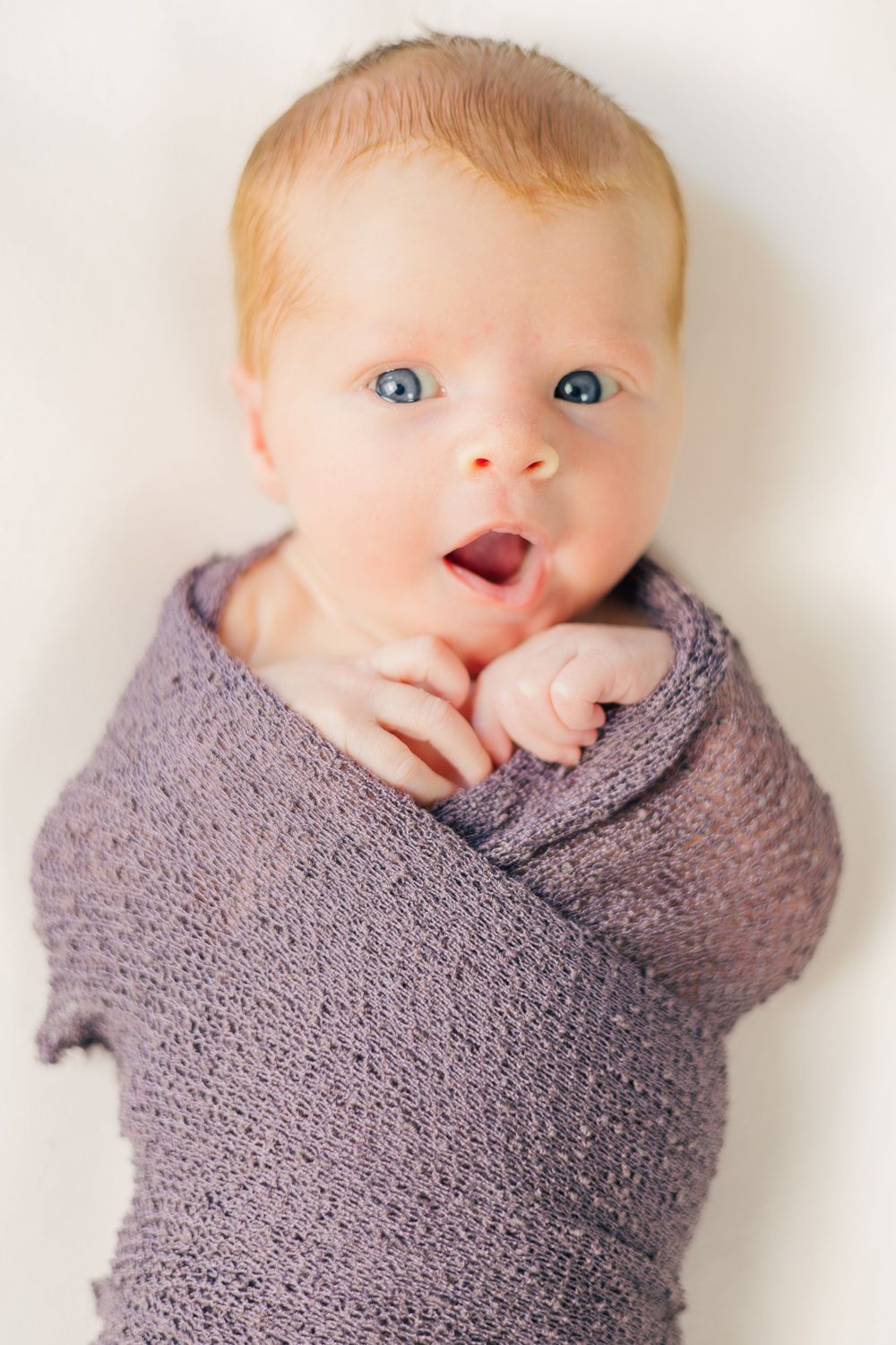 Baby swaddled in gray blanket with bright blue eyes and playful expression.