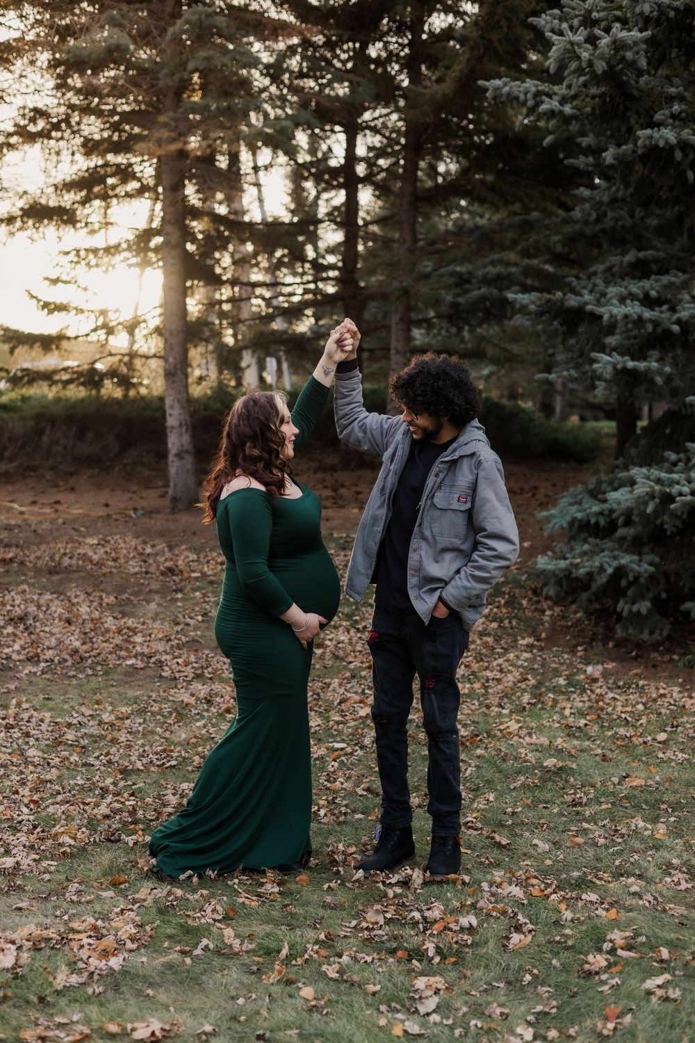 Couple sharing joyful moment during outdoor maternity photoshoot among pine trees.