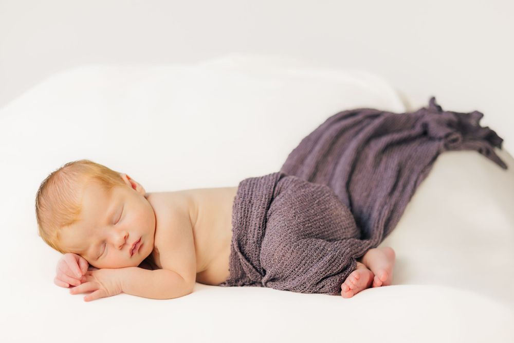 Sleeping baby wearing gray pants lying on white blanket in peaceful newborn portrait.