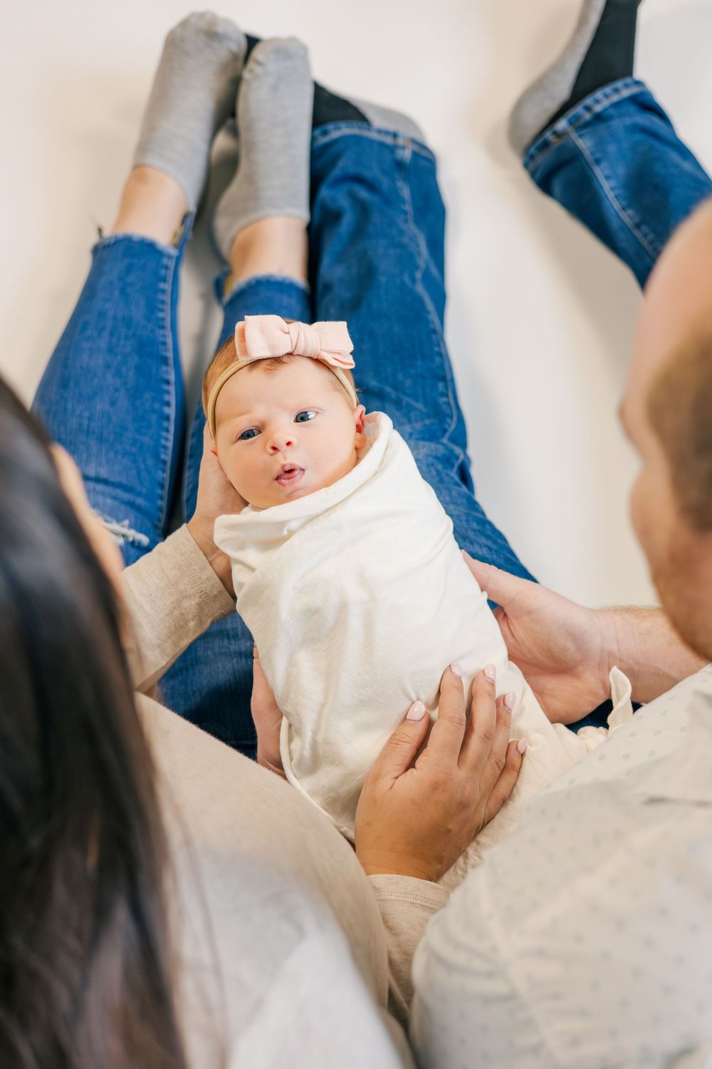 Happy baby in white onesie and bow headband laying on parents' laps.