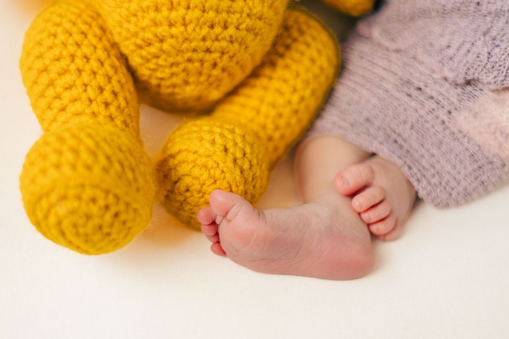 Tiny baby feet next to yellow knit stuffed toy on white background.