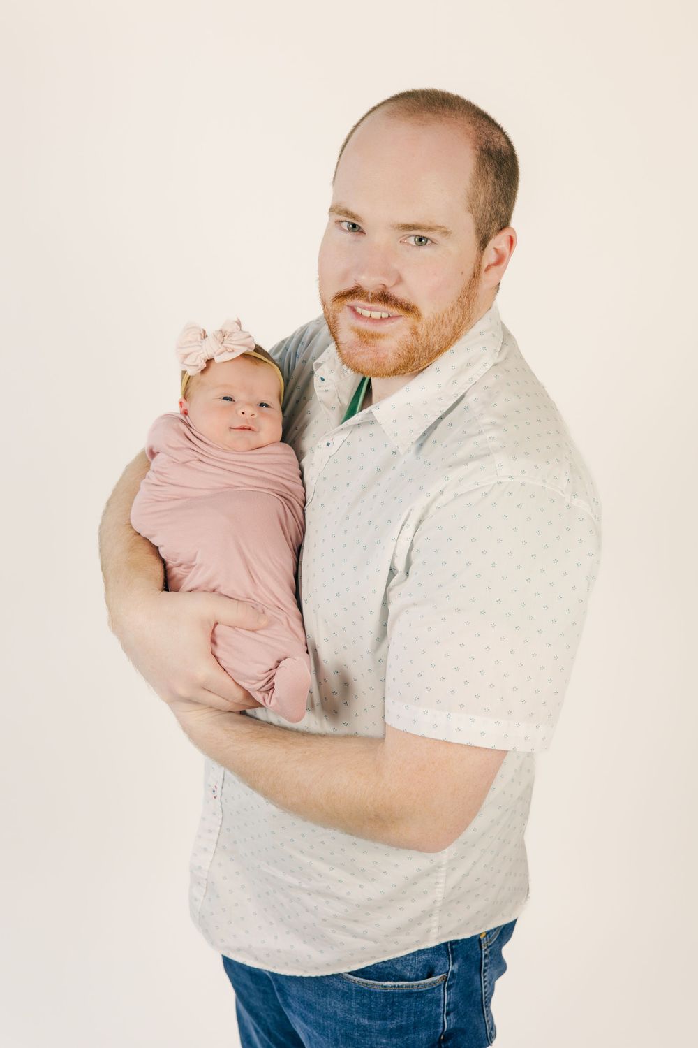 Tender moment with infant wearing pink swaddle being held against gray polka dot shirt.