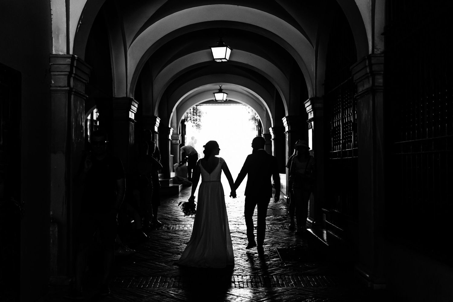 Silhouetted couple holds hands while walking through dark arched corridor in dramatic black and white photo in Seville, Spain