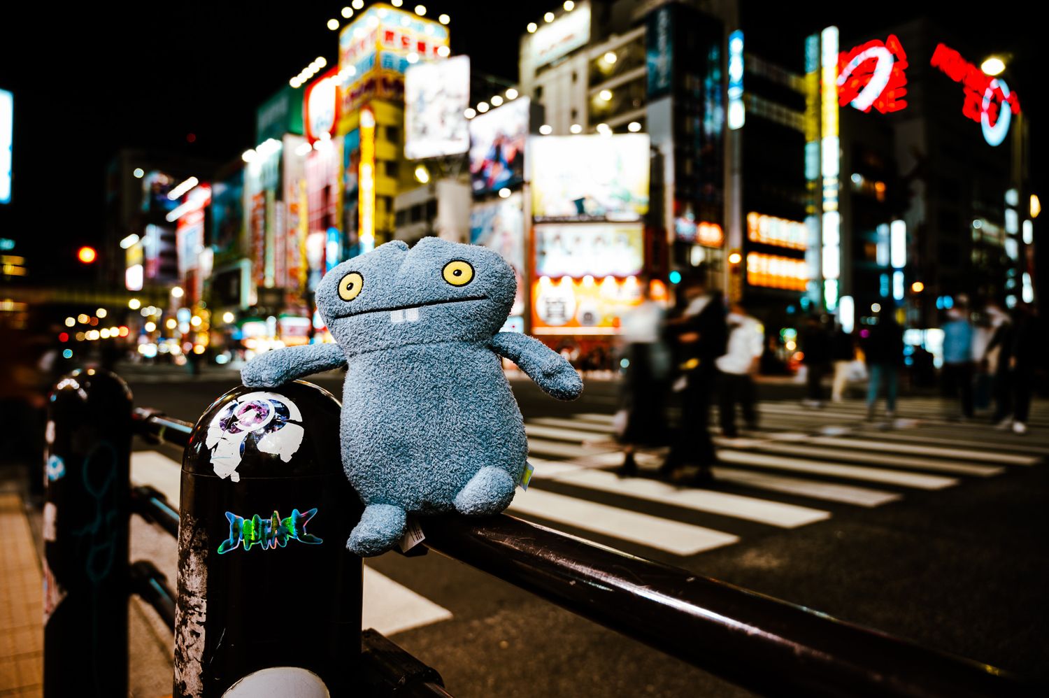 Blue plush toy sitting on railing against bright neon city lights at night.