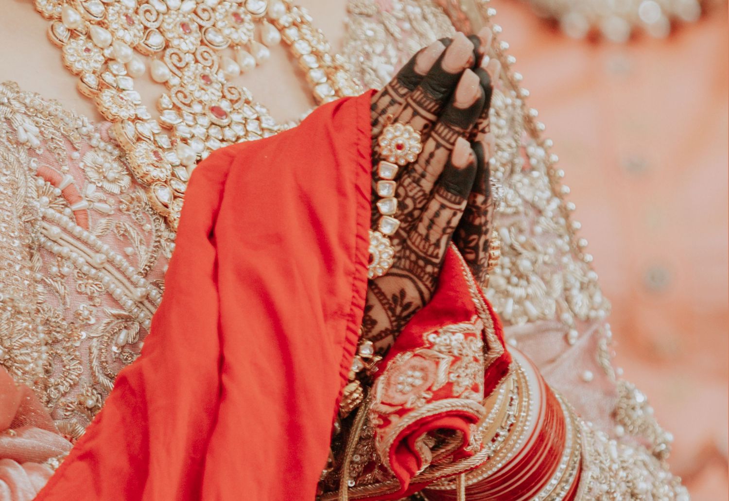 Close-up of Indian bride's hands with intricate mehndi, red bangles, and embroidered bridal outfit.