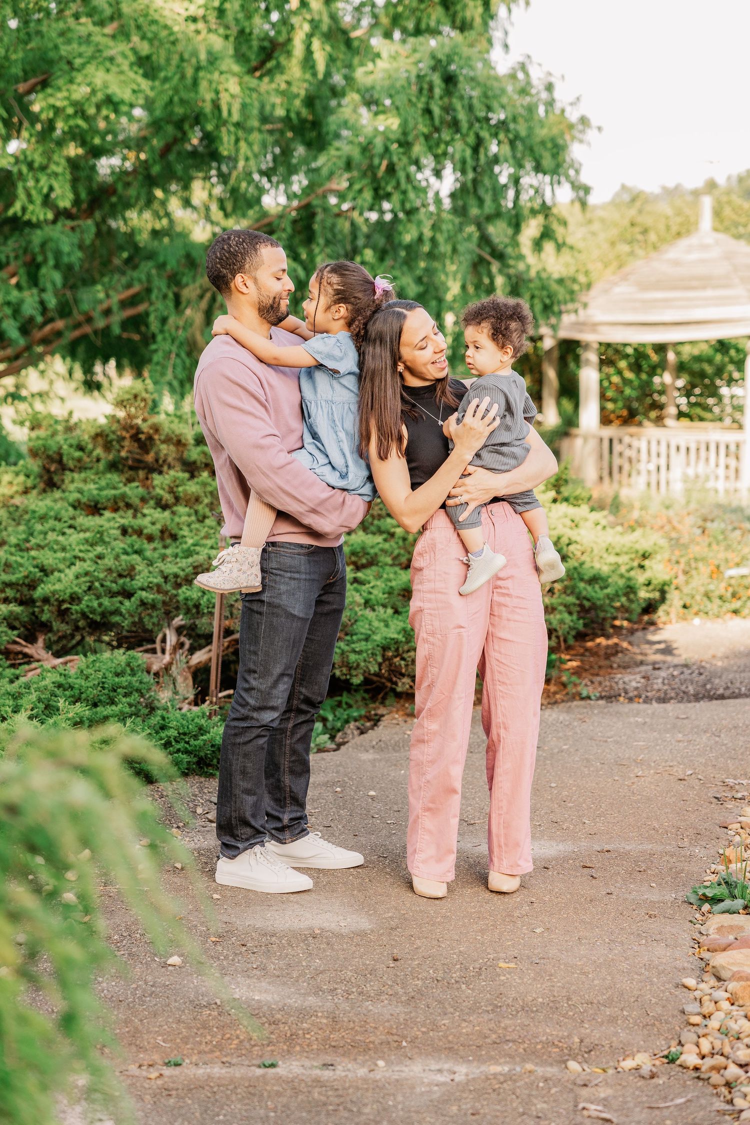 Family of four sharing an intimate moment together on a garden path with a gazebo in background.