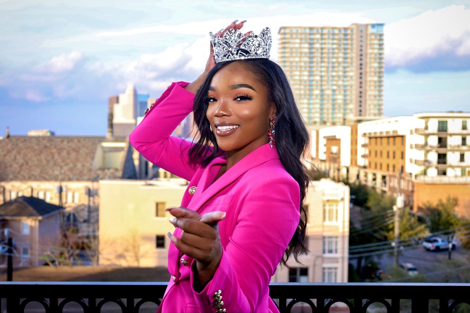 Beauty pageant winner in bright pink blazer smiling on urban balcony.