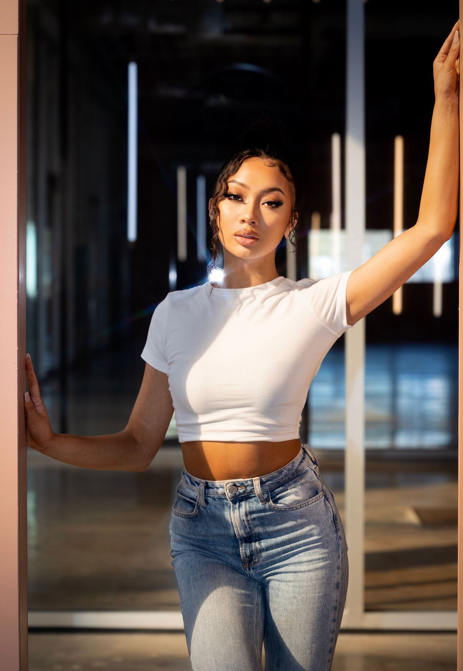 Fashion model poses in cropped white t-shirt and jeans in modern hallway with dramatic lighting.