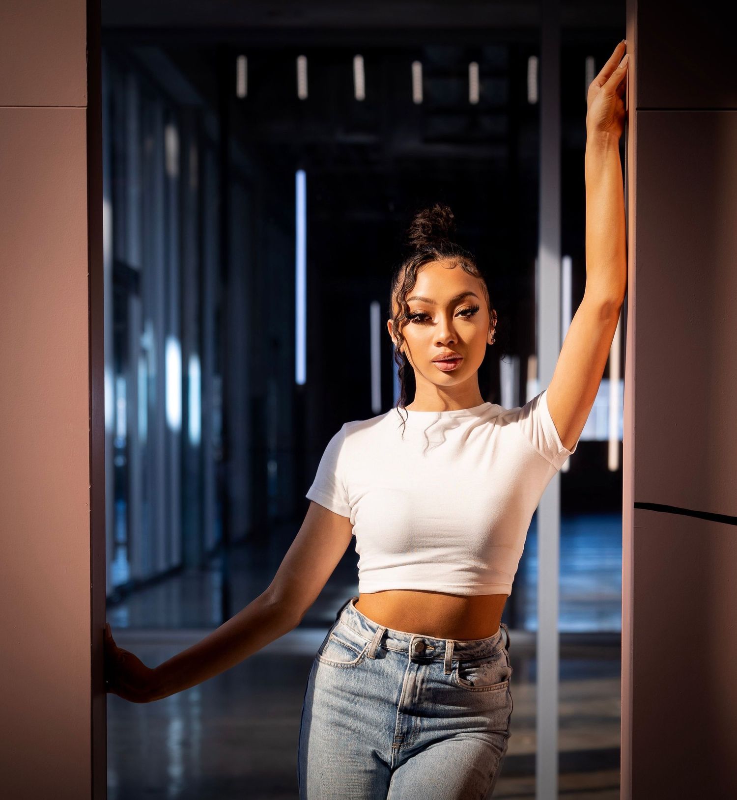 Model strikes dynamic pose in white crop top and jeans against dark interior backdrop.