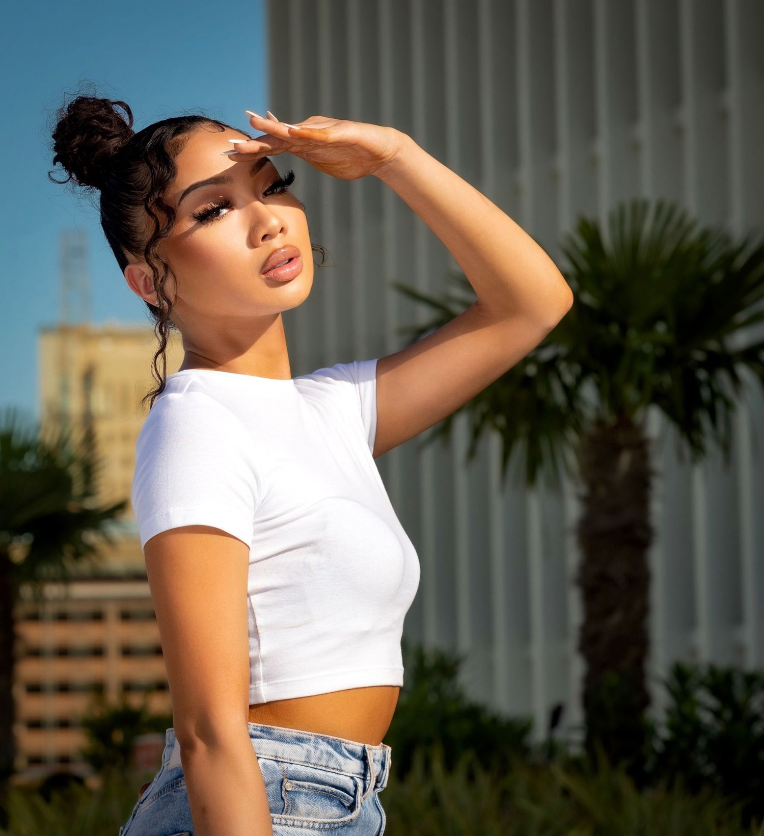 Fashion portrait of model in white crop top and jeans against urban background with palm trees.