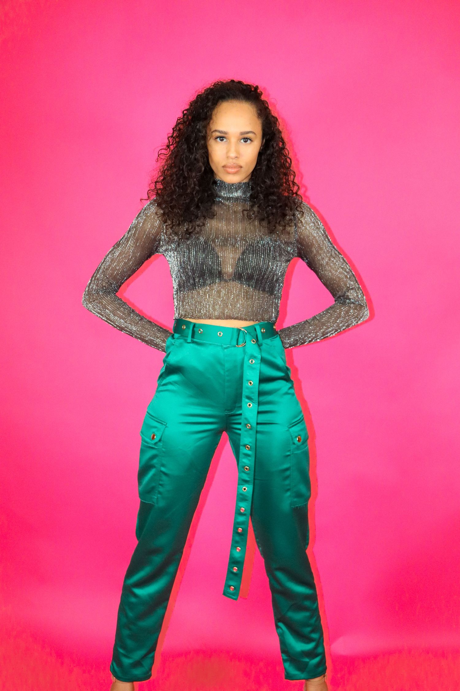 Fashion model wearing sheer mesh top and emerald green cargo pants poses against bright pink background.