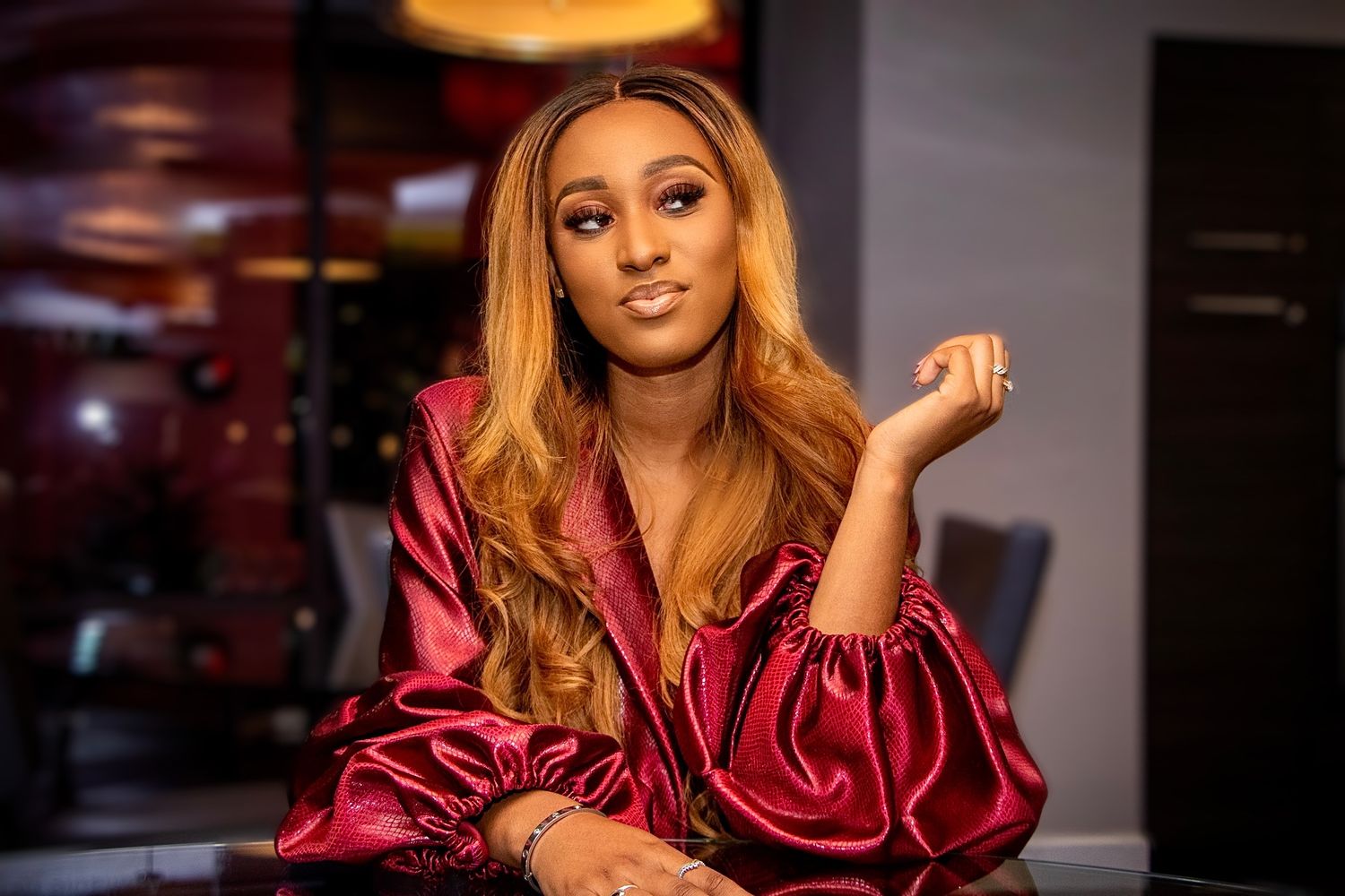 Woman with flowing caramel hair wearing red silk blouse poses playfully at elegant dining venue.