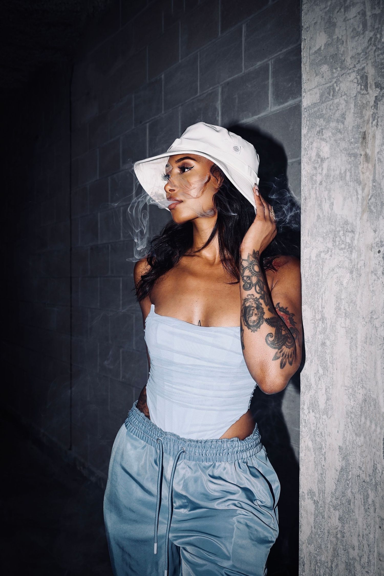 Model wearing white bucket hat and tank top with visible tattoos against dark background.