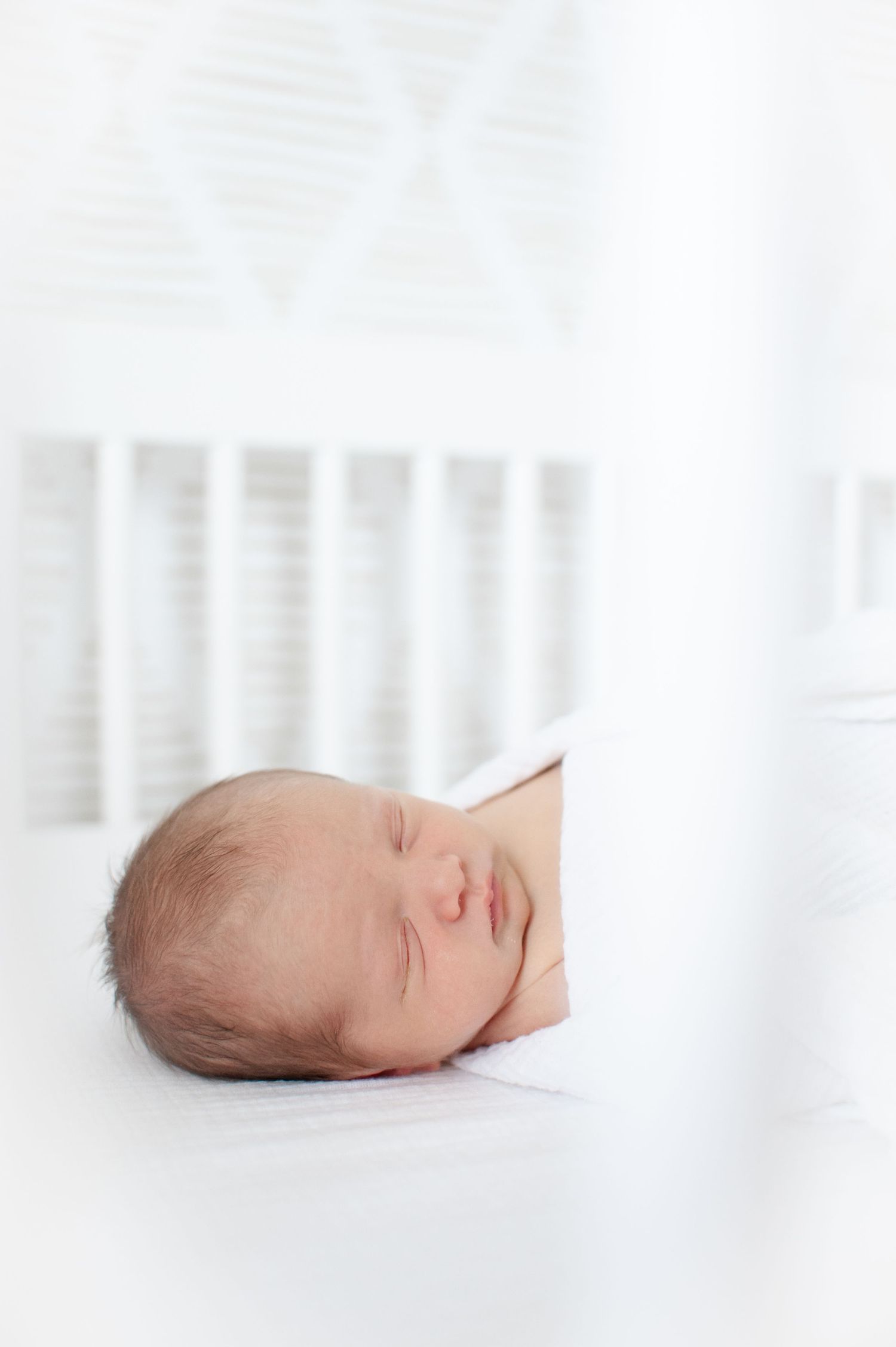 baby swaddled and sleeping in his crib.  white swaddle on white sheet with white background