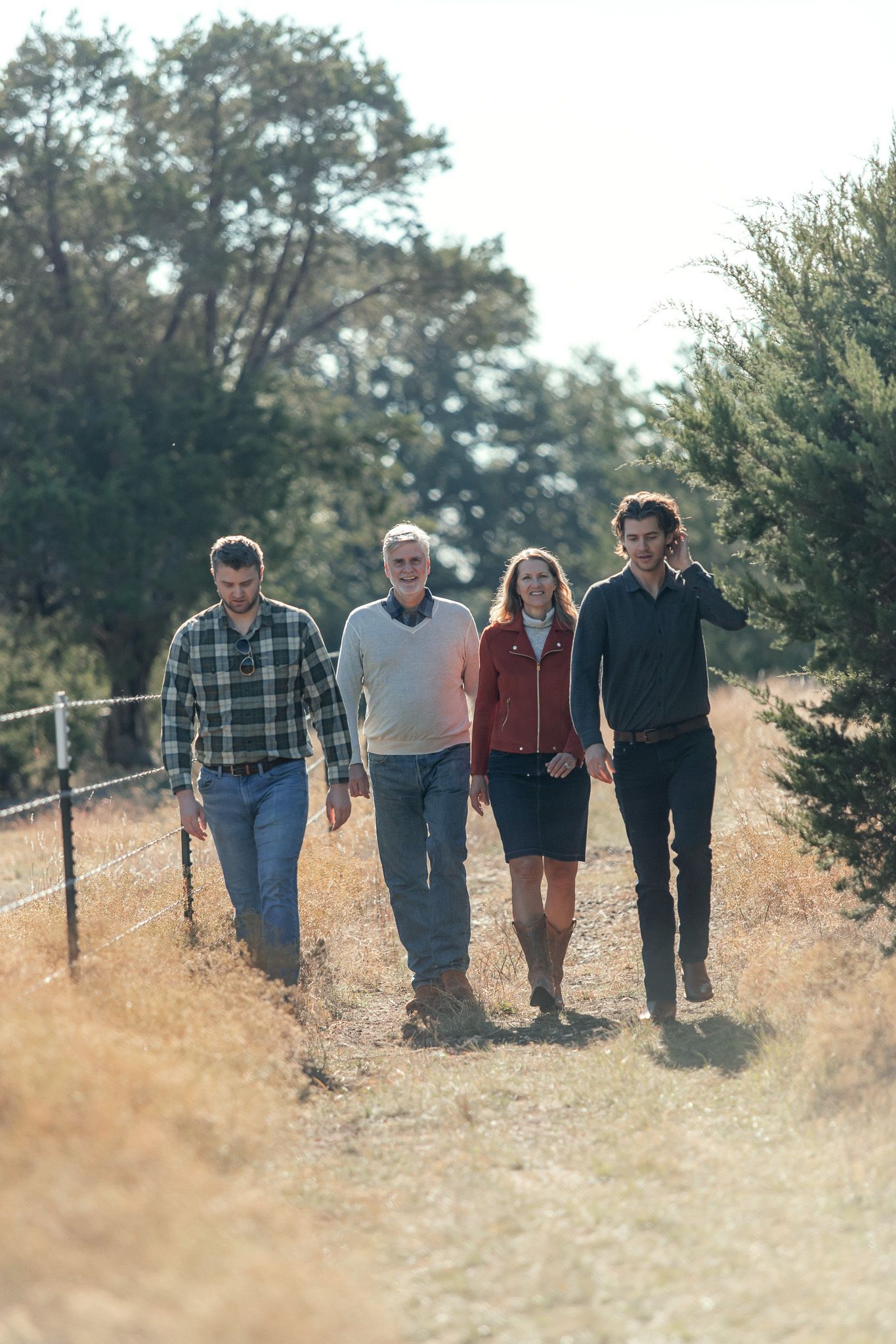 Four people walking together on a sunlit dirt path with fencing and trees.